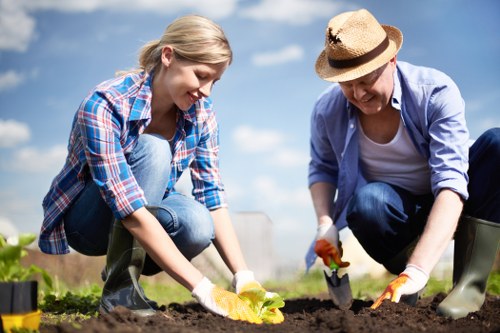 A young woman and an older woman are working together in a garden, planting or tending to soil in a landscaped outdoor space. The woman on the left, dressed in a plaid shirt and jeans, is kneeling and placing a small plant or seedling into the dark, well-tilled soil. The woman on the right, wearing a straw hat, a light blue shirt, and rubber gardening gloves, is also kneeling and assisting with the planting process. The garden area features a neatly maintained lawn in the foreground with lush green grass, and beyond the planting site, there are shrubs and trees under a partly cloudy sky, suggesting a bright, mild day ideal for outdoor gardening activities. This scene is set in a residential garden or backyard, reflecting typical gardening and landscaping work, which is relevant to the services offered by Landscapers Earls Court, specializing in outdoor space enhancement and lawn care in the Earl's Court area near London.
