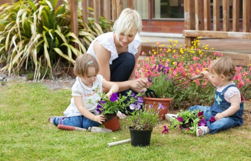 A smiling woman with short blonde hair is sitting on a well-maintained lawn in a backyard, engaging with two young children who are planting flowers in small pots. The woman wears a white top and dark pants, while the children, a girl and a boy, are dressed in casual summer clothes; the girl in a white t-shirt with a floral pattern and the boy in a blue t-shirt with denim shorts. They are surrounded by colourful potted plants, including purple and pink flowers, with a backdrop of a wooden deck, a variety of lush green plants, and a flower bed with yellow and pink blossoms. The garden appears tidy and thoughtfully landscaped, featuring a mix of grass, soil, and flower beds, with natural light suggesting a bright and pleasant day. The scene emphasizes outdoor gardening activities typical of professional landscaping and garden maintenance services, highlighting a family-friendly outdoor space in the Earls Court area, suitable for outdoor leisure and gardening projects managed by Landscapers Earls Court.