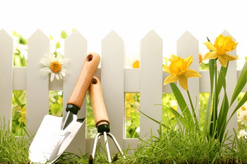 The image depicts a small garden scene with a white picket fence in the background, partially shaded by sunlight. In the foreground, there is well-maintained green grass with a dense, lush texture. To the right, vibrant yellow daffodils with long green stems and leaves are blooming beside the fence. To the left, a single white daisy with a yellow centre is visible, also growing near the fence. Resting at the base of the fence, there are two gardening tools: a hand trowel with a wooden handle and a garden fork with a metal head, both placed on the grass, suggesting recent or upcoming garden maintenance. The overall environment appears bright with natural daylight, creating a fresh and inviting outdoor setting typical of a well-kept garden space in London or nearby areas, suitable for gardening and outdoor landscape enhancement by local professionals like Landscapers Earls Court.